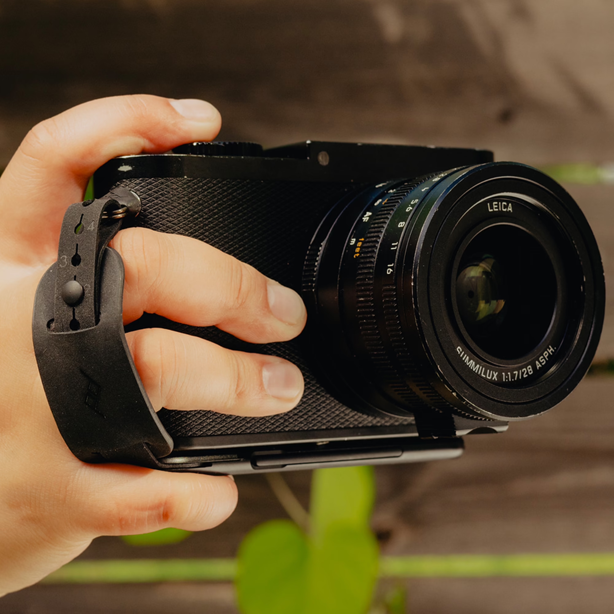 Eine Hand hält eine schwarze Leica Kamera mit montierter Peak Design Micro Clutch Handschlaufe in der Praxisanwendung vor einem unscharfen, natürlichen Hintergrund im Freien.
