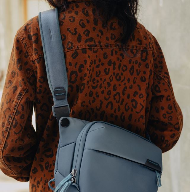 Rückansicht einer Person mit einer braunen Jacke im Leopardenmuster, die eine taubenblaue Peak Design Everyday Sling V2 Kameratasche (in der Farbvariante Ocean) cross-body diagonal über den Rücken trägt. Das Lifestyle-Foto fokussiert auf das ergonomisch g