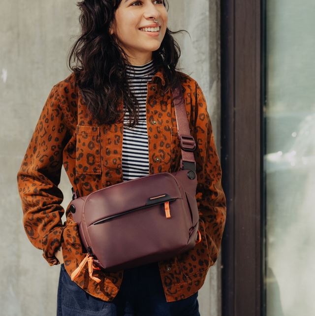 Eine lächelnde Frau im Halbprofil von vorne, die eine weinrote Peak Design Everyday Sling V2 Kameratasche (in der Farbe Eclipse) cross-body an einem farblich abgestimmten Schultergurt trägt. Die wetterfeste Umhängetasche schmiegt sich an den Oberkörper an