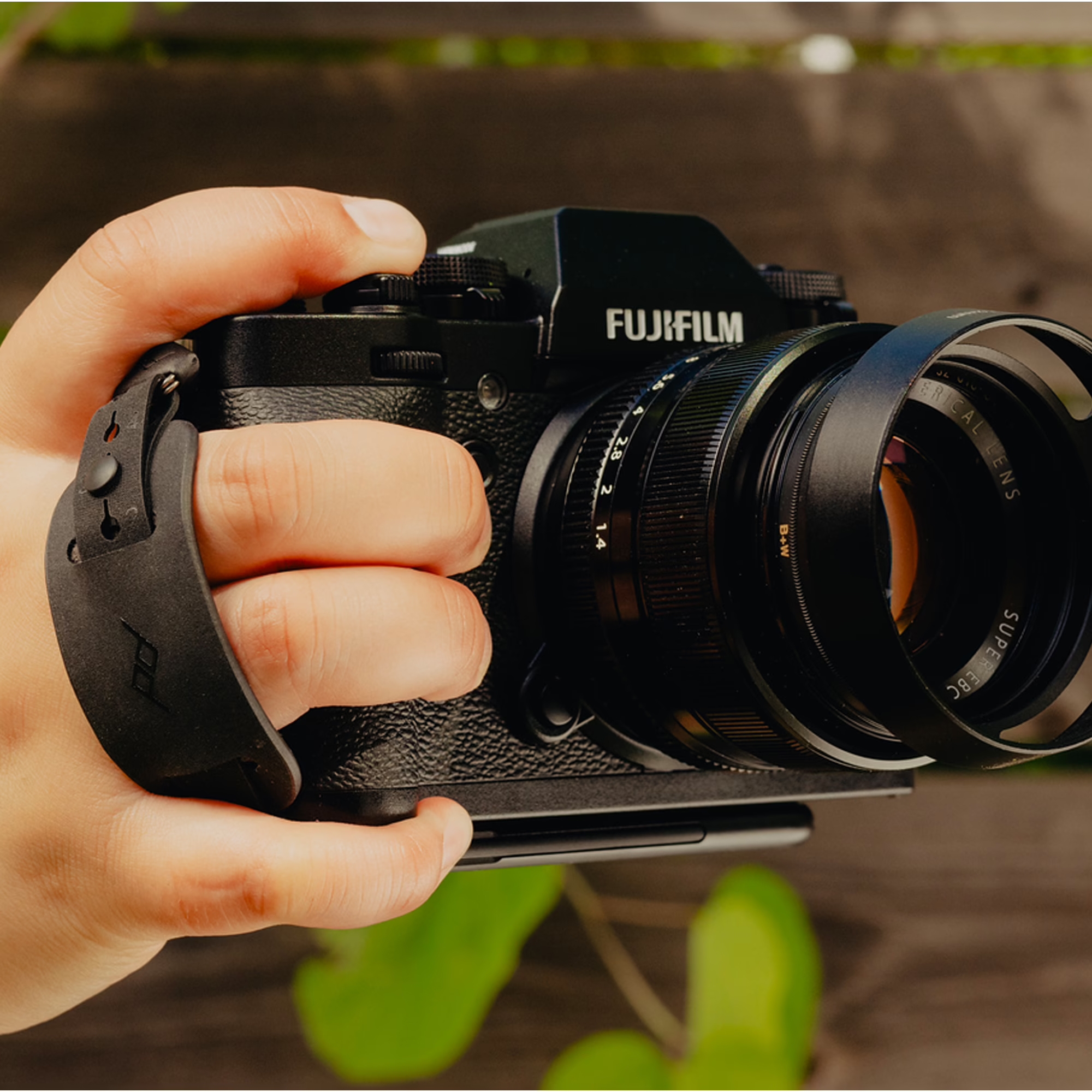Nahaufnahme einer Hand, die eine schwarze Fujifilm Systemkamera mit angesetztem Objektiv und montierter Peak Design Micro Clutch Handschlaufe hält.